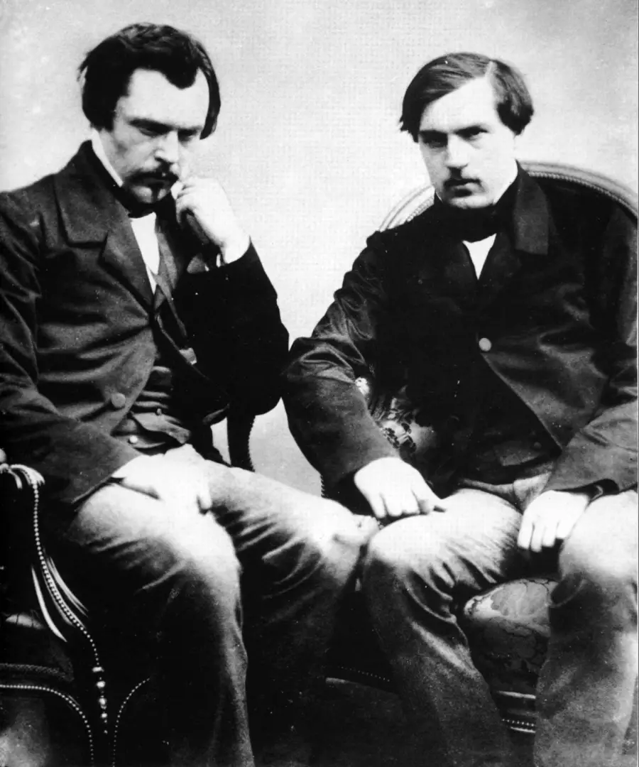 Two men in 19th-century clothing sit side by side on ornate chairs, both looking seriously at the camera. One man rests his chin on his hand in a thoughtful pose, while the other sits with his hands on his knees.