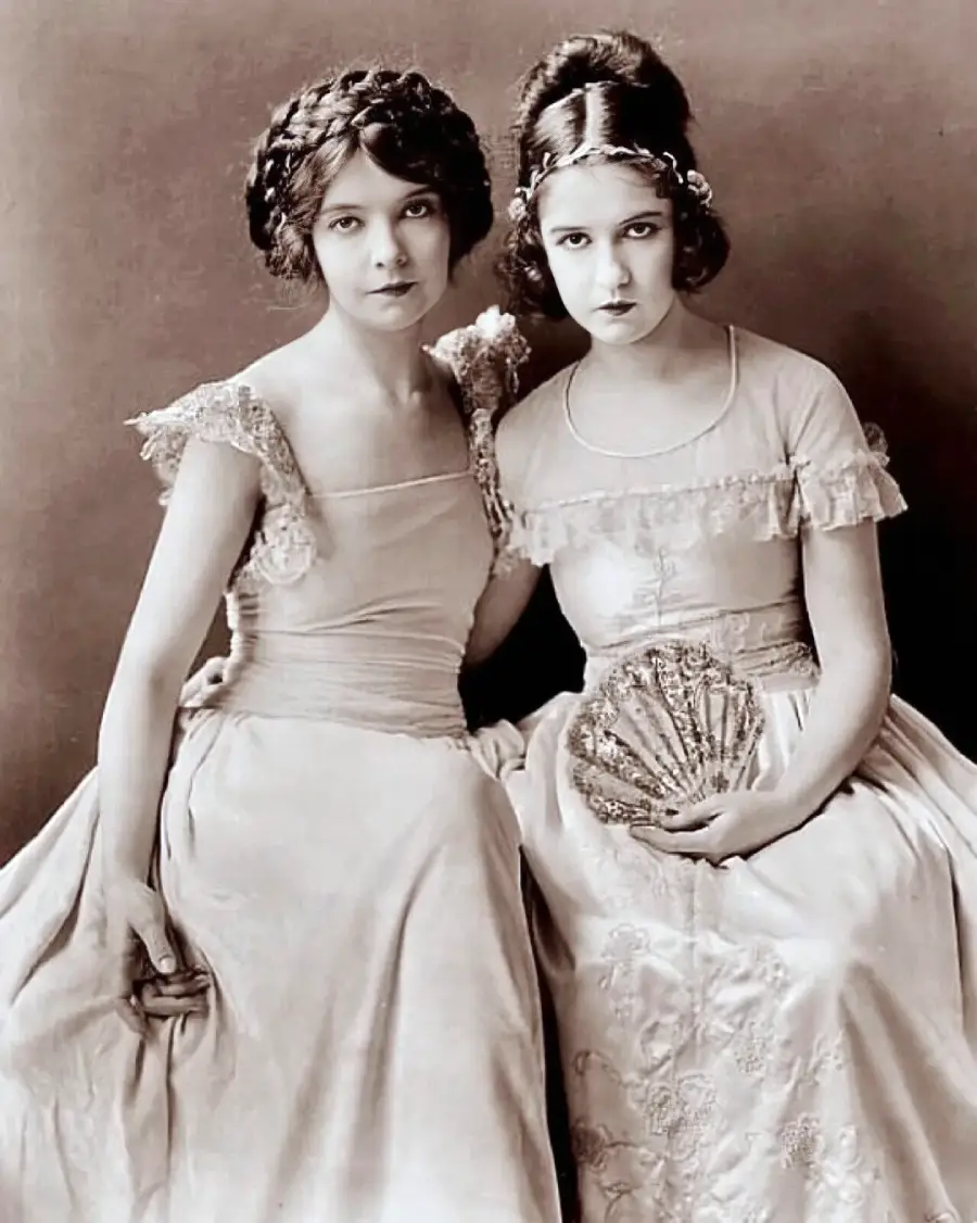 Two young women in vintage formal dresses sit closely together, looking at the camera. One holds an ornate fan. Their hairstyles and attire suggest the early 20th century. The photo is in black and white.