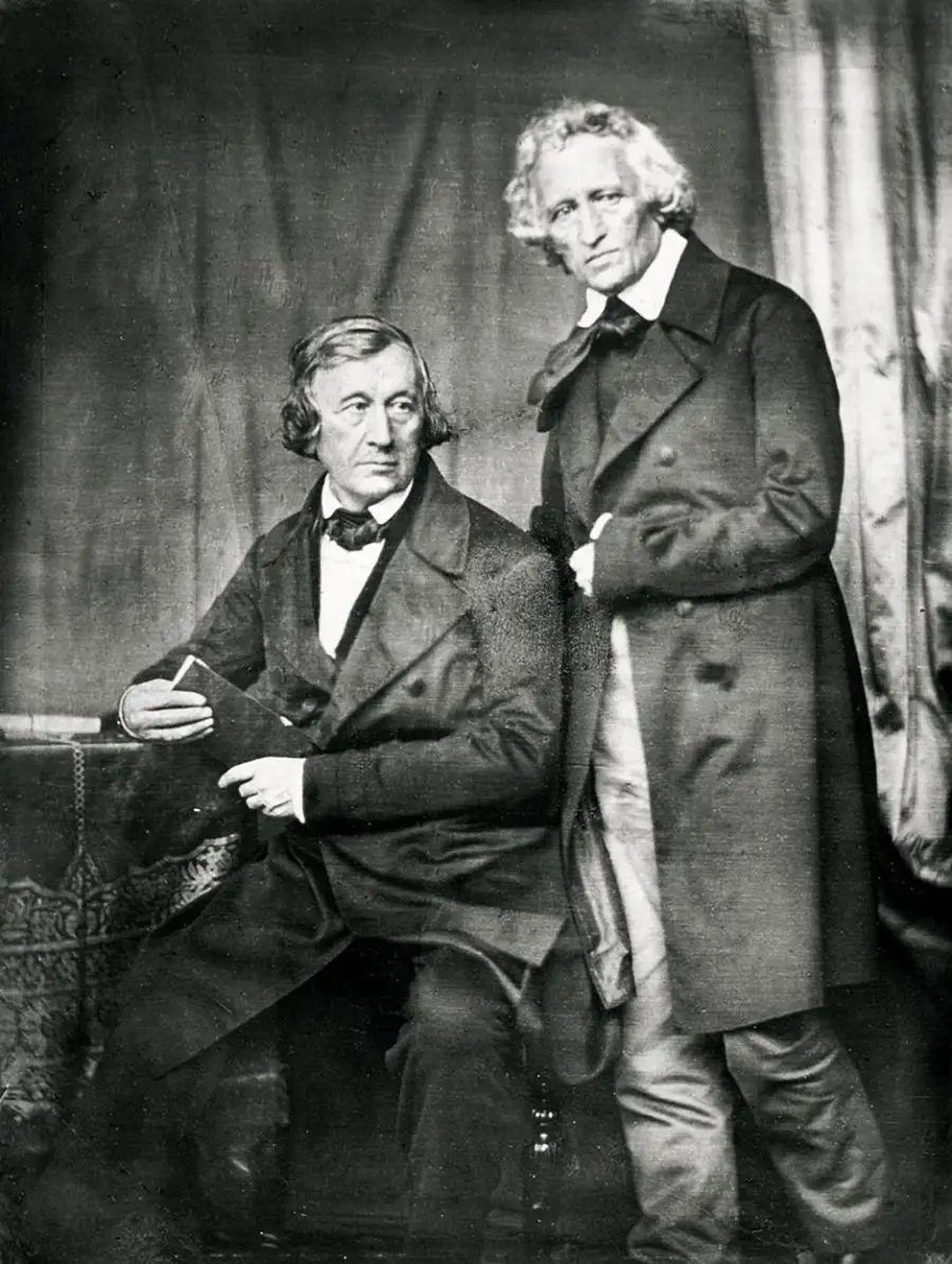 Two older men with wavy hair and serious expressions wear dark 19th-century formal coats. One is seated, holding a book, while the other stands beside him, both posing for a vintage black-and-white photograph.