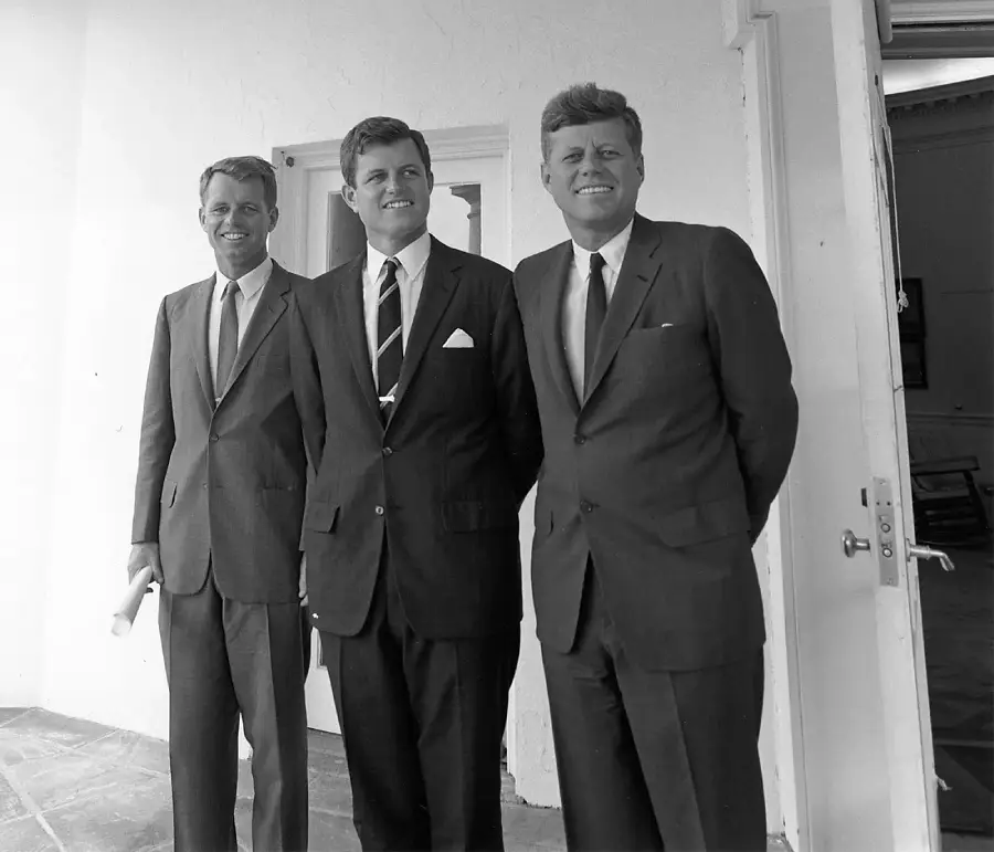 Three men in suits stand side by side, smiling, in front of a light-colored building with an open door. Two have their hands by their sides, and one holds a rolled document.
