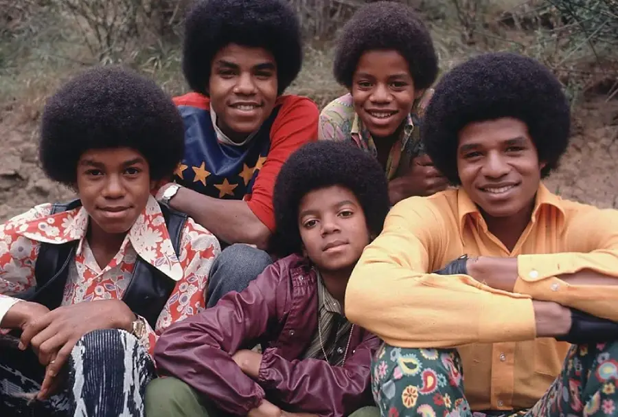 Five young boys with afros sit outdoors, smiling at the camera. They wear colorful, patterned 1970s clothing, including shirts with bold prints and bright colors. Greenery is visible in the background.