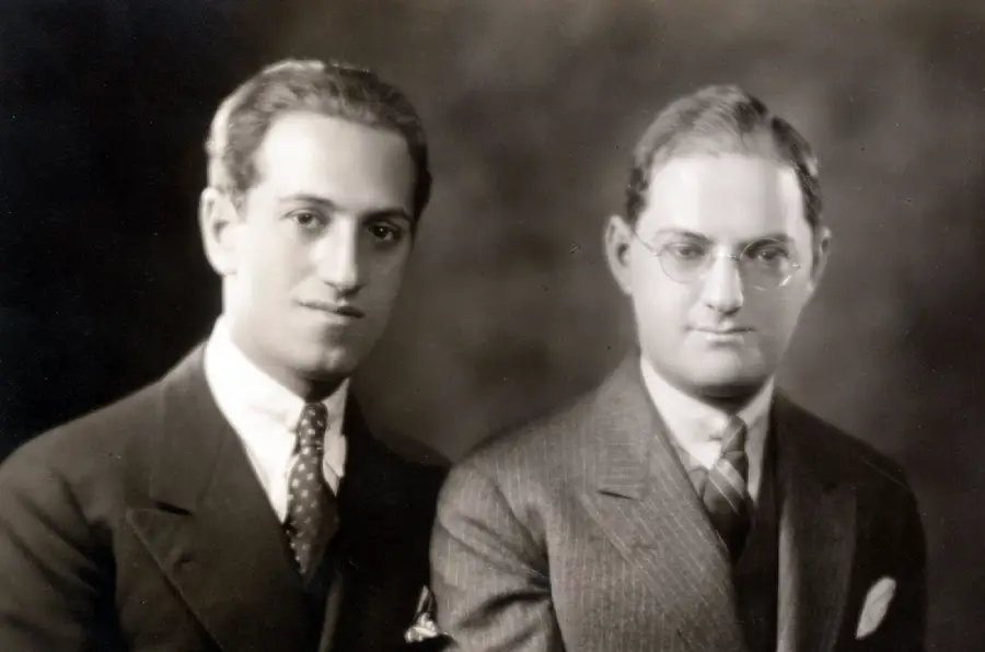 Two men in suits pose for a formal black-and-white portrait against a neutral background. One man has dark hair and no glasses; the other has lighter hair and wears round glasses.