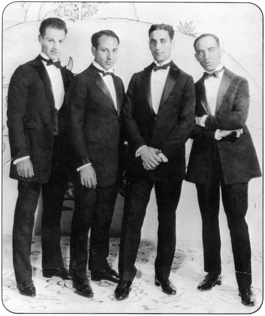 Four men dressed in formal tuxedos stand side by side, posing for a black-and-white portrait in front of a decorated backdrop. They appear confident and composed, with hands at their sides or arms crossed.