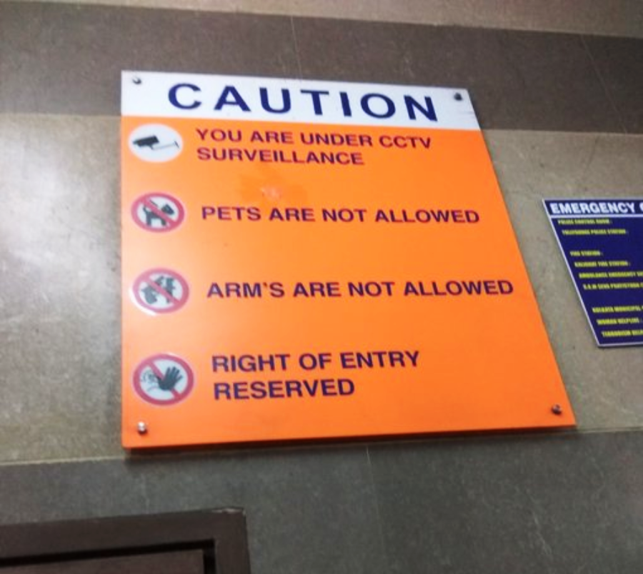 An orange caution sign on a wall states: "You are under CCTV surveillance, Pets are not allowed, Arm's are not allowed, Right of entry reserved," with icons for each rule. A blue emergency contact sign is nearby.