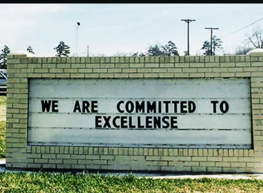 A brick sign with black letters reads, "WE ARE COMMITTED TO EXCELLENSE," with a humorous misspelling of "excellence." The sign is outdoors, with grass and trees in the background.