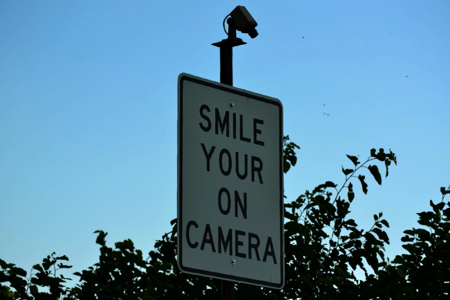 A security camera is mounted above a sign that reads “SMILE YOUR ON CAMERA,” with a grammatical error. Green foliage and a clear blue sky are visible in the background.