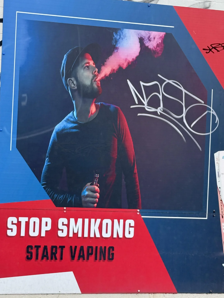 A billboard shows a man exhaling vapor with the text "STOP SMIKONG START VAPING." The sign is defaced with graffiti and the image is set on a blue and red background.