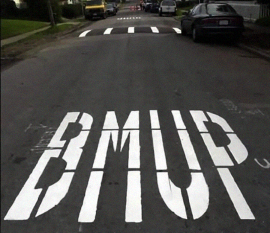 Street with large, white painted letters spelling "BMUP" instead of "BUMP," warning drivers of a speed bump ahead; cars are parked along the sides and a yellow speed bump is visible in the distance.