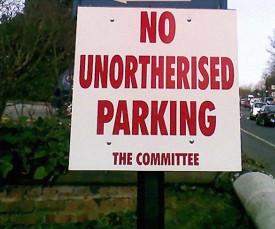 A sign by the roadside reads "NO UNORTHERISED PARKING THE COMMITTEE" in bold red letters, with a spelling error in the word "UNOTHERISED." Shrubs and cars are visible in the background.