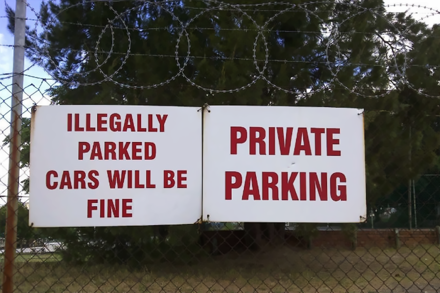 Two large white signs on a chain-link fence topped with barbed wire. One sign reads "ILLEGALLY PARKED CARS WILL BE FINE" and the other reads "PRIVATE PARKING," both in bold red letters. Trees are visible behind the fence.