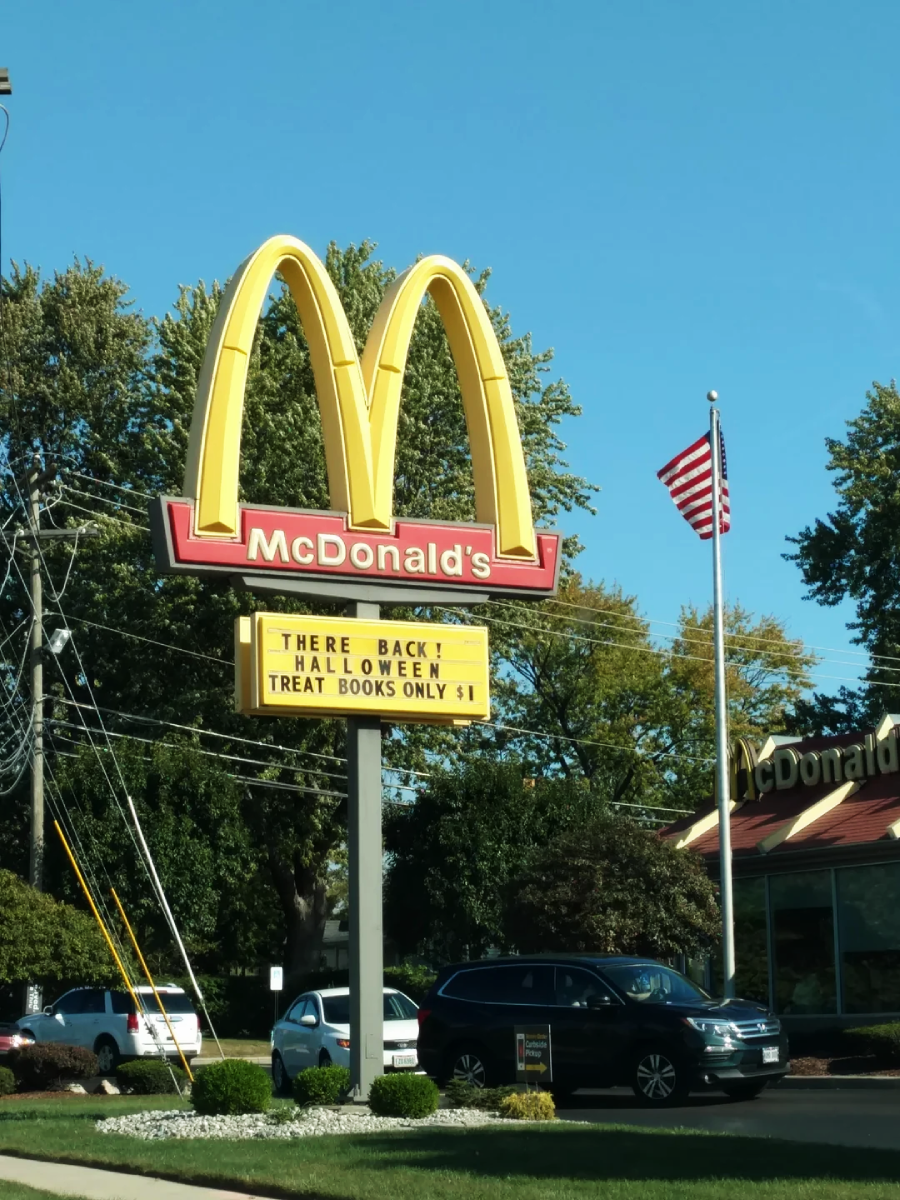 A McDonald’s restaurant sign with a message board that reads, "THEY'RE BACK HALLOWEEN TREAT BOOKS ONLY $1." An American flag and several cars are visible in the background.