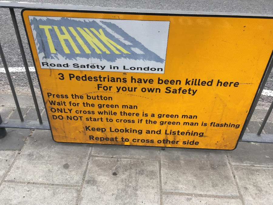 A yellow road safety sign in London warns that three pedestrians have been killed here. It instructs to press the button, wait for the green man, and only cross when safe, urging people to stay alert.