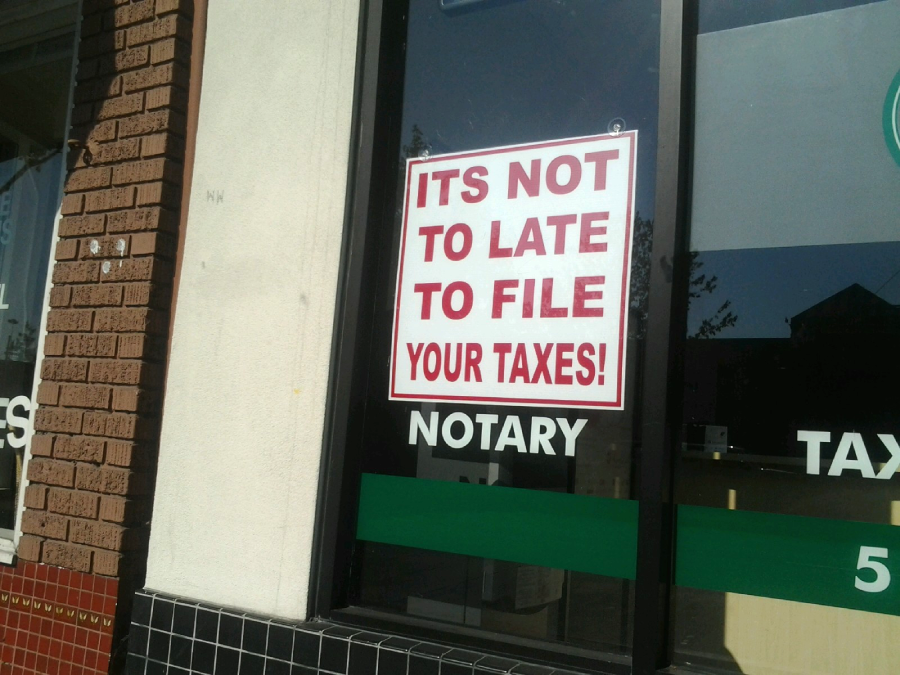 A sign in a window reads, "ITS NOT TO LATE TO FILE YOUR TAXES!" in bold red letters. The sign is taped to the glass of a notary office.