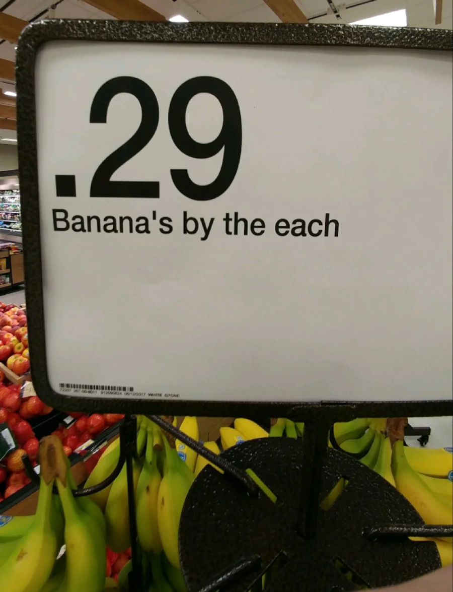 A grocery store sign reads "0.29 Banana's by the each" with bananas displayed below and apples in the background. The sign contains a grammatical error, using "Banana's" instead of "Bananas.
