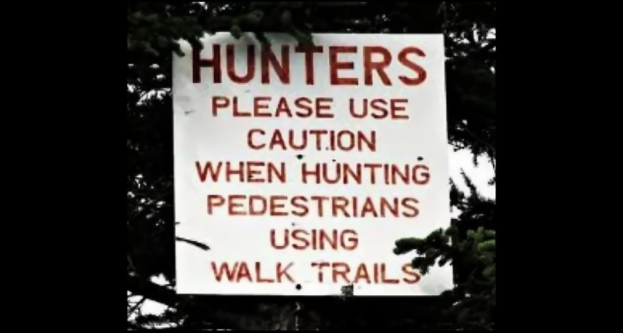 A sign reads: "HUNTERS PLEASE USE CAUTION WHEN HUNTING PEDESTRIANS USING WALK TRAILS," with the text in red and surrounded by tree branches.