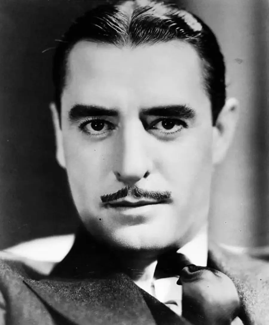 Black and white close-up portrait of a man with slicked-back hair, a thin mustache, and a serious expression, wearing a suit and holding a pipe near his chin.