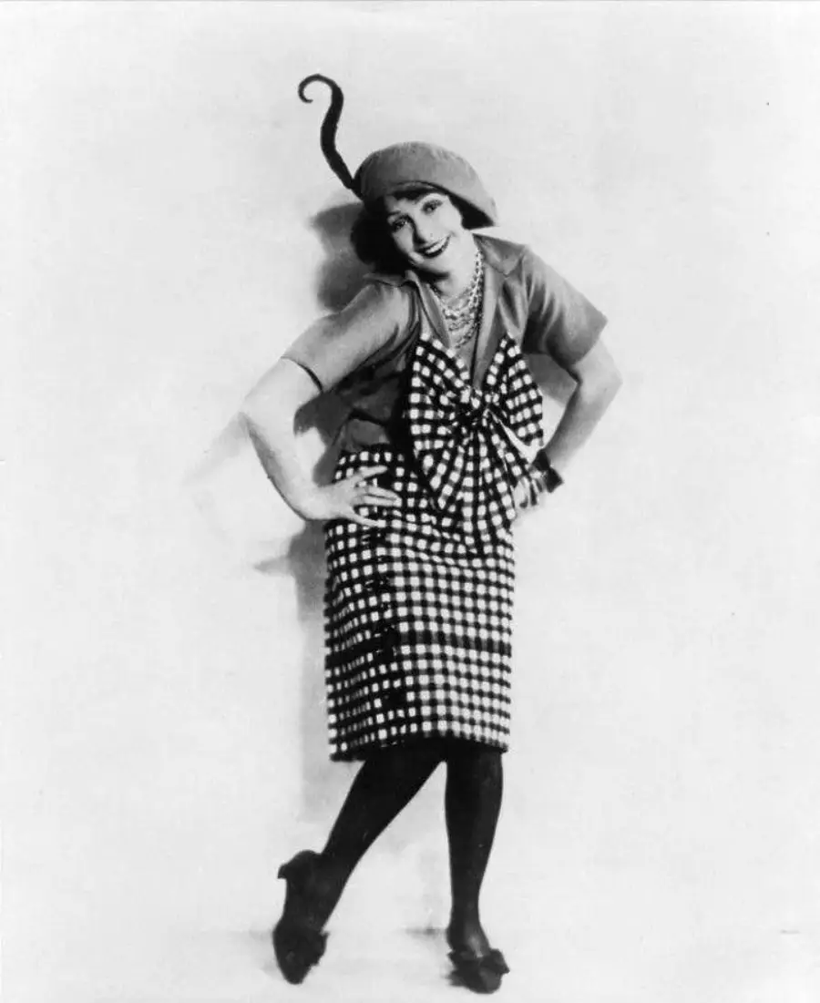 A woman poses playfully, wearing a checkered skirt, a matching large bow on her top, dark stockings, and a whimsical hat with a curled antenna-like extension. The photo is black and white.