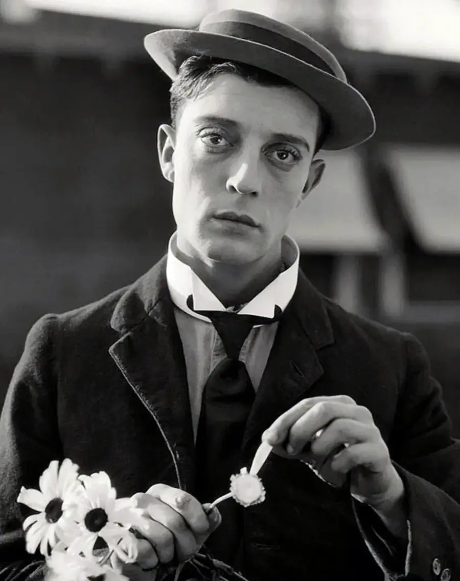 A man in vintage clothing and a tilted hat holds a bouquet of daisies and plucks petals from one flower, looking somberly at the camera. The image is in black and white.