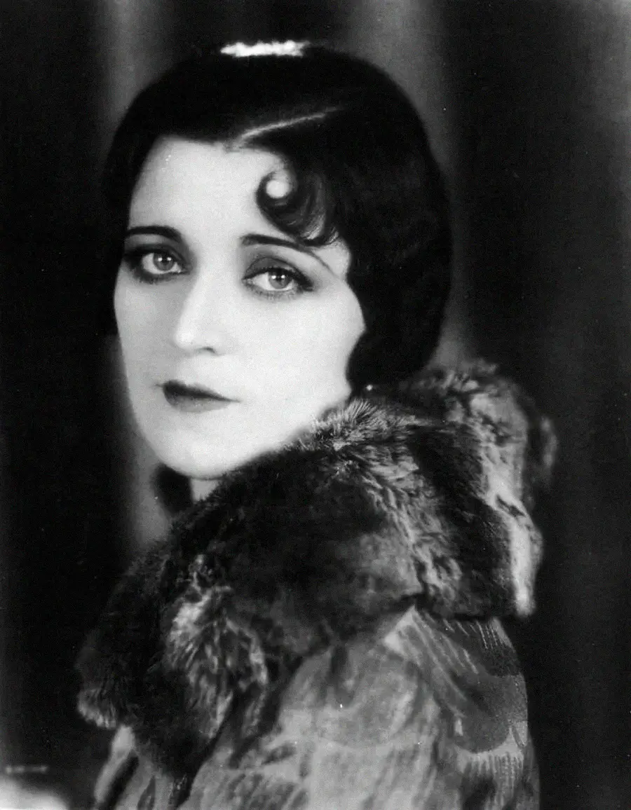 A black-and-white portrait of a woman with short, wavy dark hair, styled in the 1920s fashion. She is wearing a coat with a fur collar and gazes softly at the camera with expressive eyes.