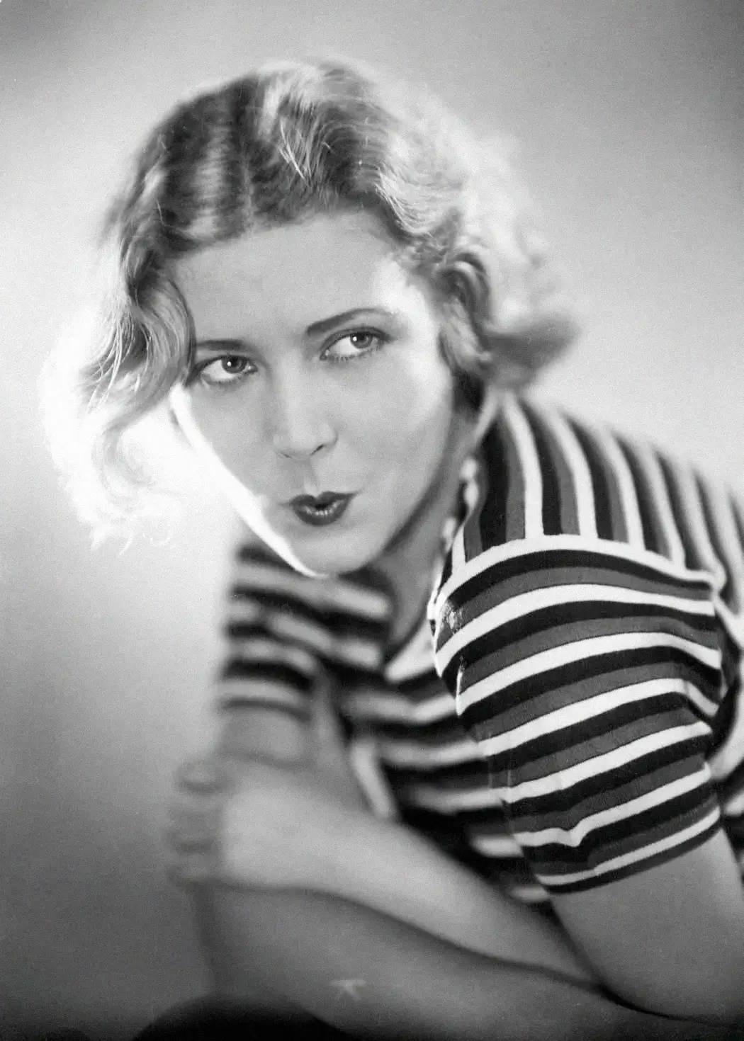 Black and white portrait of a woman with wavy, shoulder-length hair, wearing a striped shirt. She is leaning forward, looking slightly up, with pursed lips as if whistling or playfully posing.