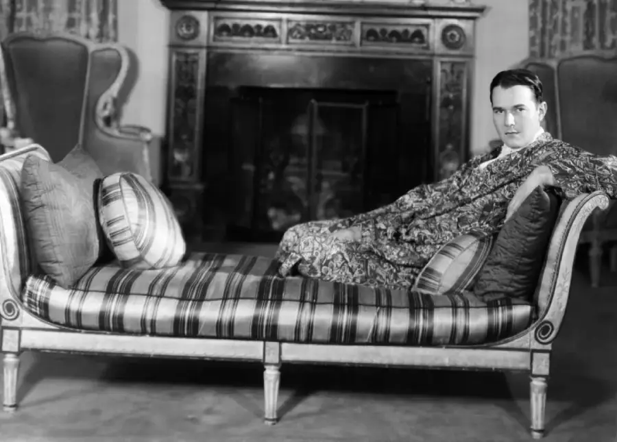A man in patterned robes reclines on a striped chaise lounge in an elegant room with ornate chairs and a decorative fireplace in the background.