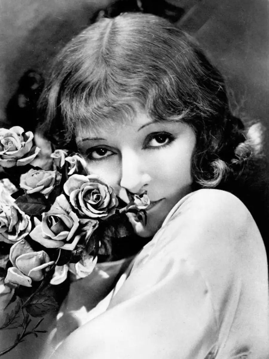Black and white photo of a woman with wavy hair holding a bouquet of roses close to her face, gazing softly at the camera.