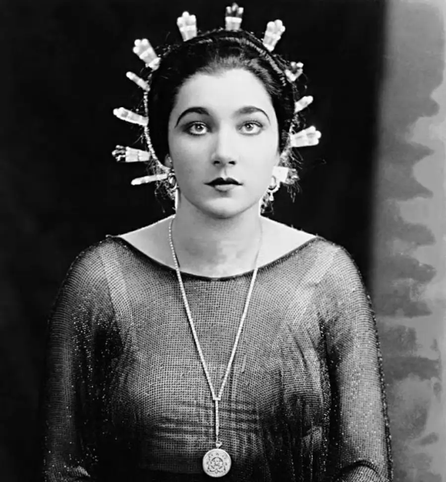 A woman in a sheer, dark top sits facing the camera, wearing a headpiece with multiple upright rods or tubes. She has dark hair pulled back and wears a long necklace with a large circular pendant. The background is dark and plain.