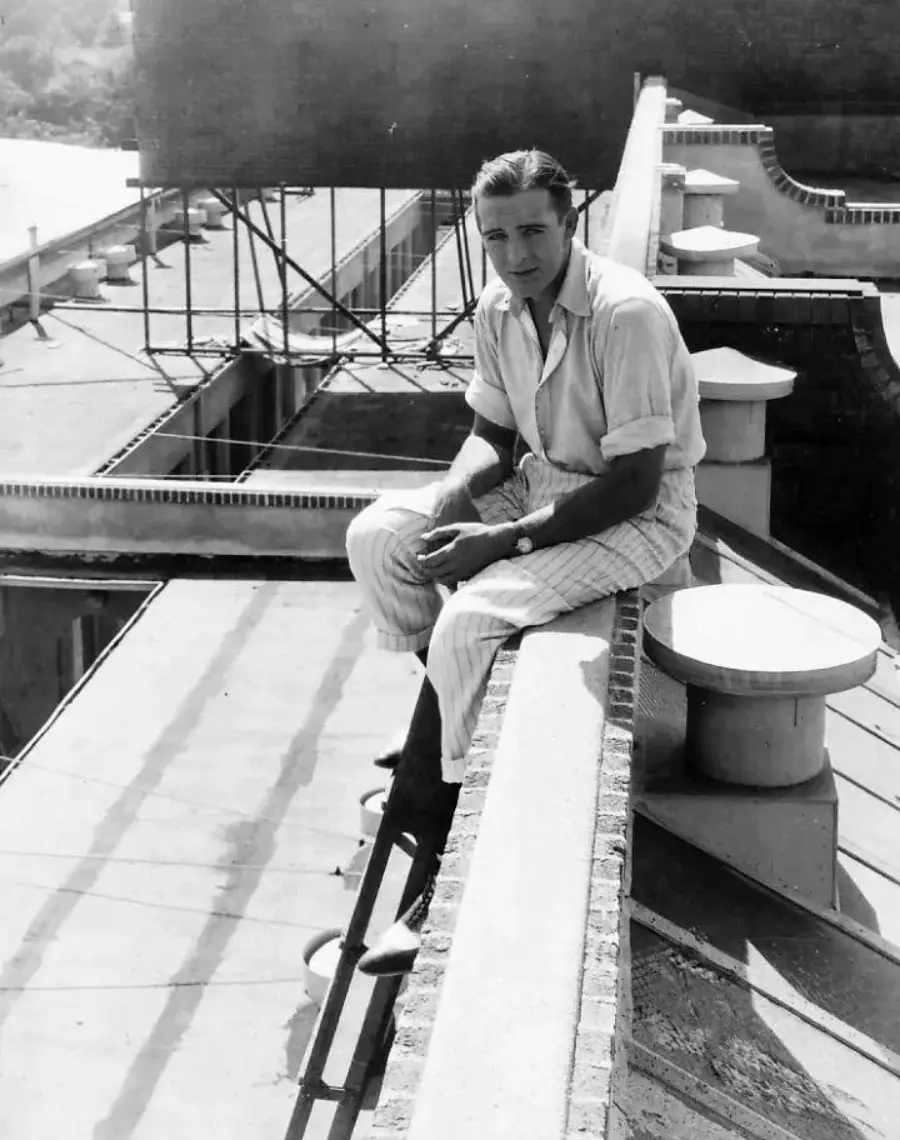 A man in a light shirt and striped pants sits on the edge of a rooftop, looking at the camera. The rooftop has brickwork and ventilation structures, with buildings and trees visible in the background.