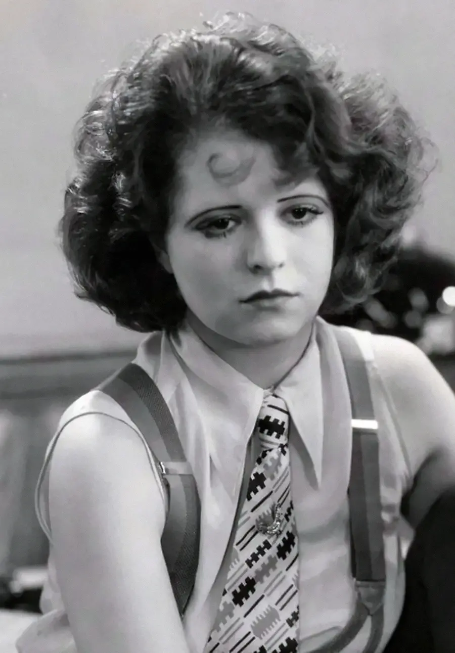 A young woman with wavy, short hair wears a patterned tie, collared shirt, and suspenders. She looks down with a serious, contemplative expression in this black and white photograph.