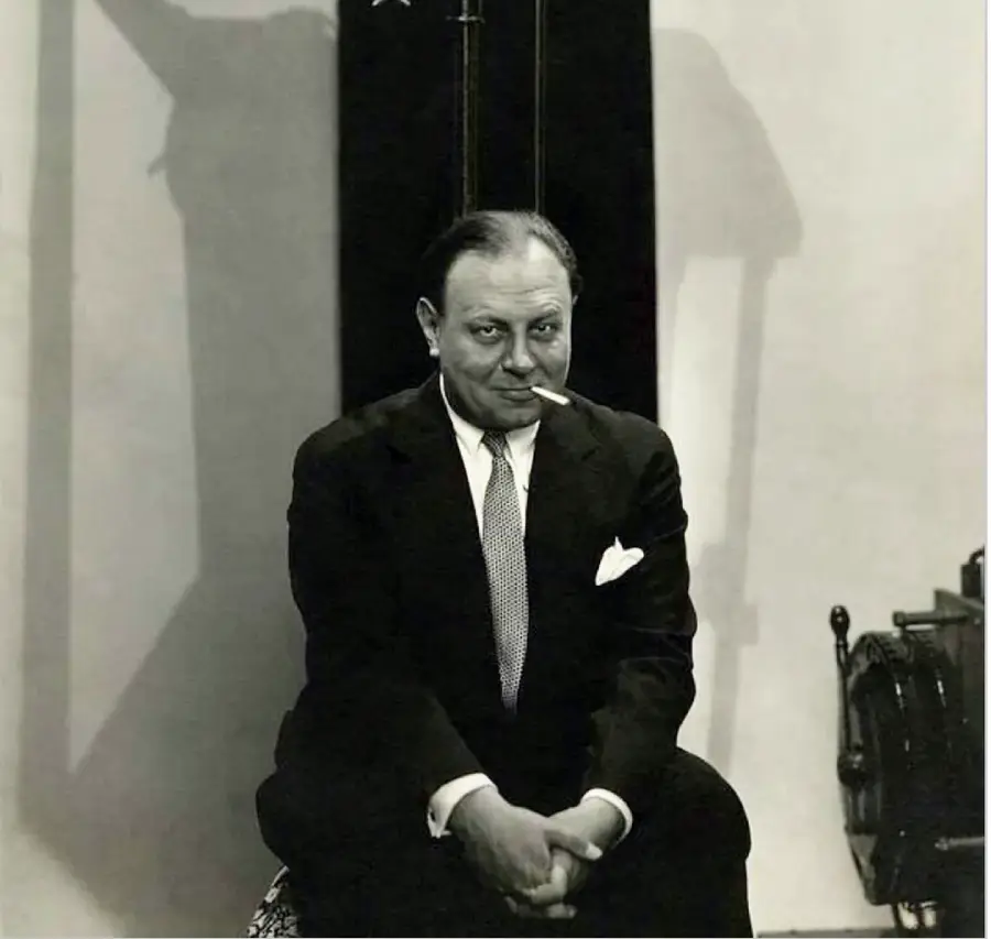 A man in a suit sits with hands clasped, looking forward with a slight smile and a cigarette in his mouth. The background is plain with a tall, dark object and a chair partially visible on the right.