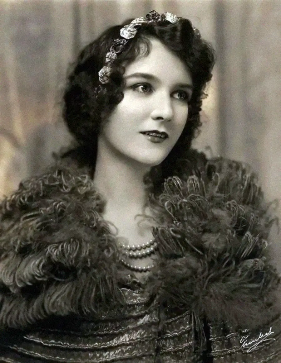 A vintage black-and-white portrait of a young woman with wavy dark hair, wearing a decorative headband, layered pearl necklace, and a textured, feathery shawl, gazing slightly to the side.