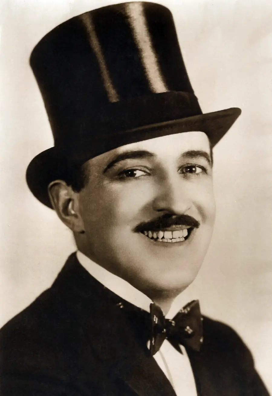 A smiling man in a tuxedo, bow tie, and tall top hat poses for a vintage black-and-white portrait.