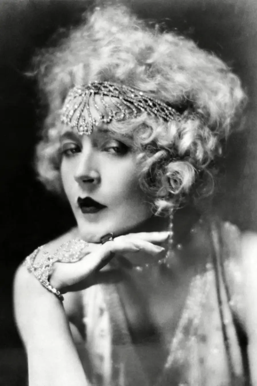 A woman with curly blonde hair wearing a jeweled headpiece and bracelet rests her chin on her hand, gazing confidently. The image is in black and white and has a vintage, glamorous feel.