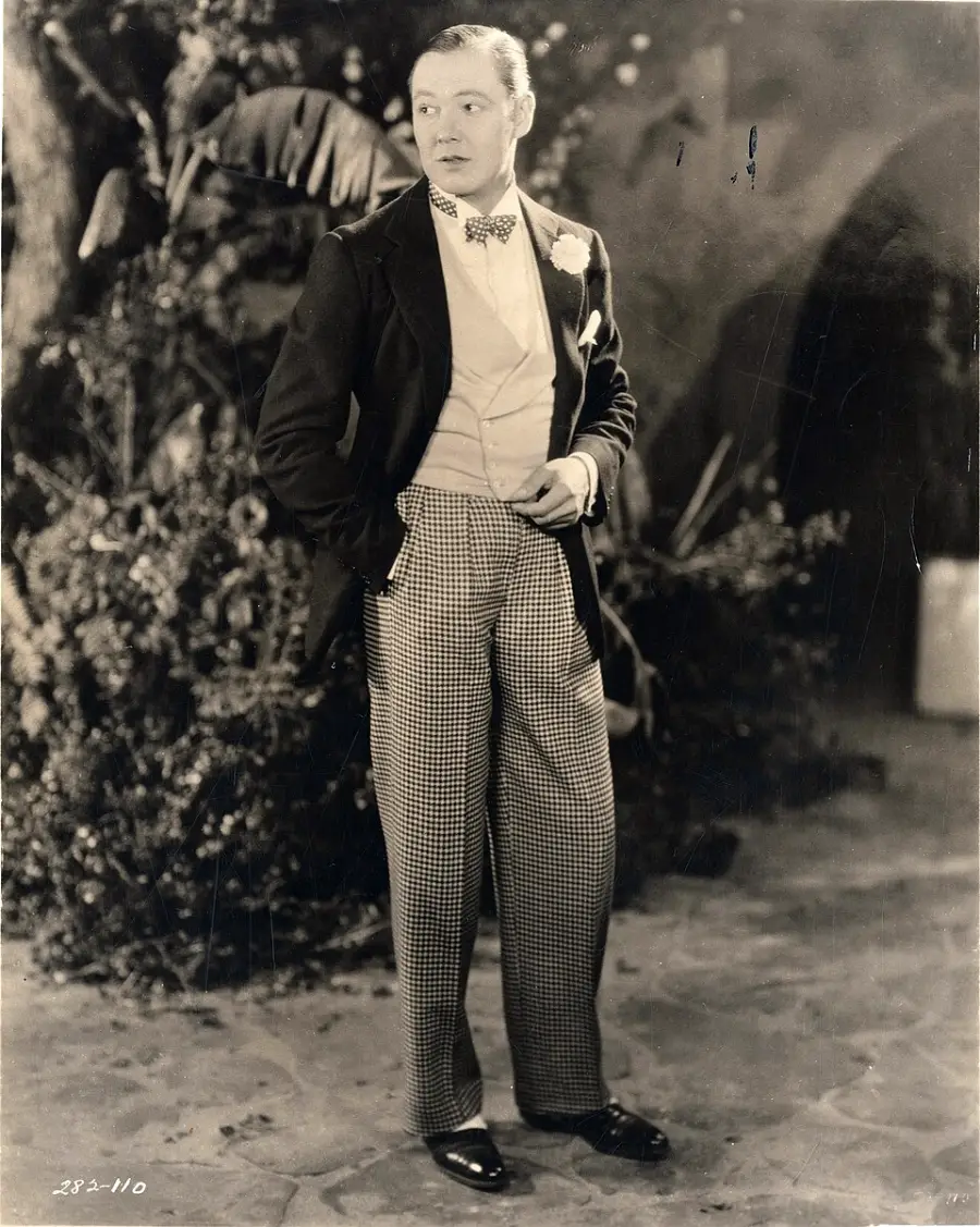 A man in formal 1920s attire stands outdoors on a stone path, wearing a tuxedo jacket, bow tie, vest, and checkered pleated trousers, with his hands in his pockets and plants in the background.