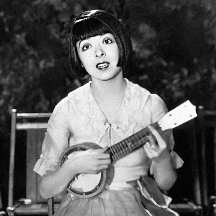 A woman with short dark hair and bangs sits outdoors playing a small banjo. She wears a light blouse and skirt, looking upward with an expressive face. The image is in black and white.