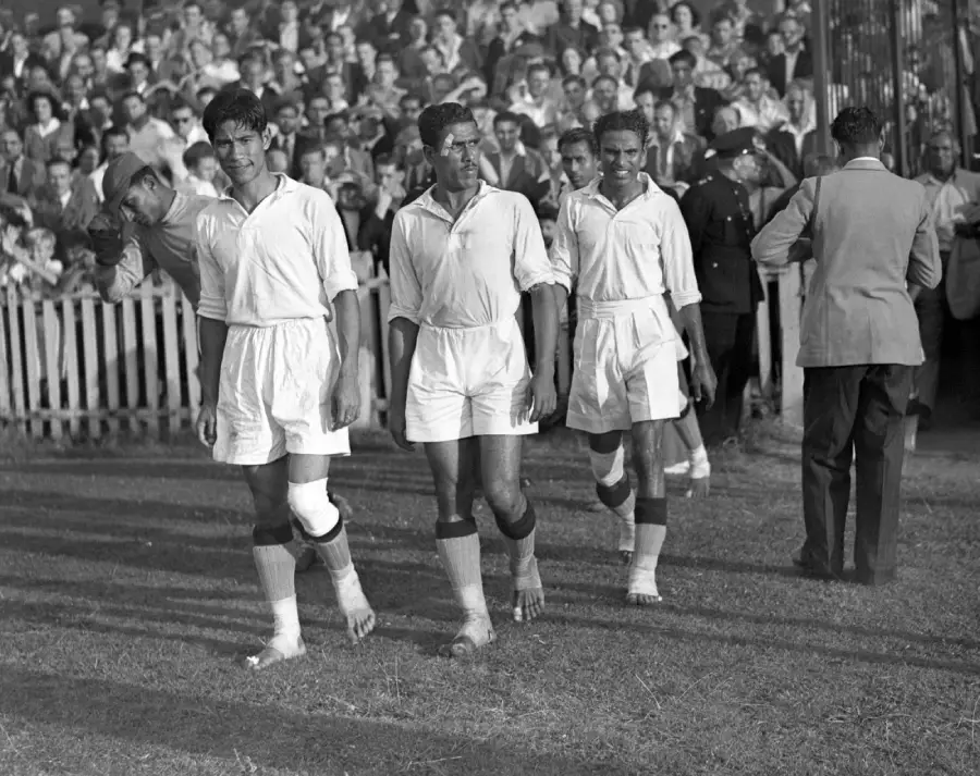 Four barefoot soccer players in white uniforms walk off a grassy field, with a crowd of spectators in the background watching from behind a fence. One player has bandaged legs. The scene appears to be from a past era.