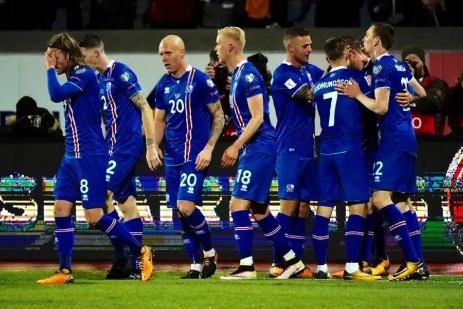 A group of soccer players in blue Iceland uniforms celebrate together on the field, some hugging while others stand nearby; one player covers his face with his hand.