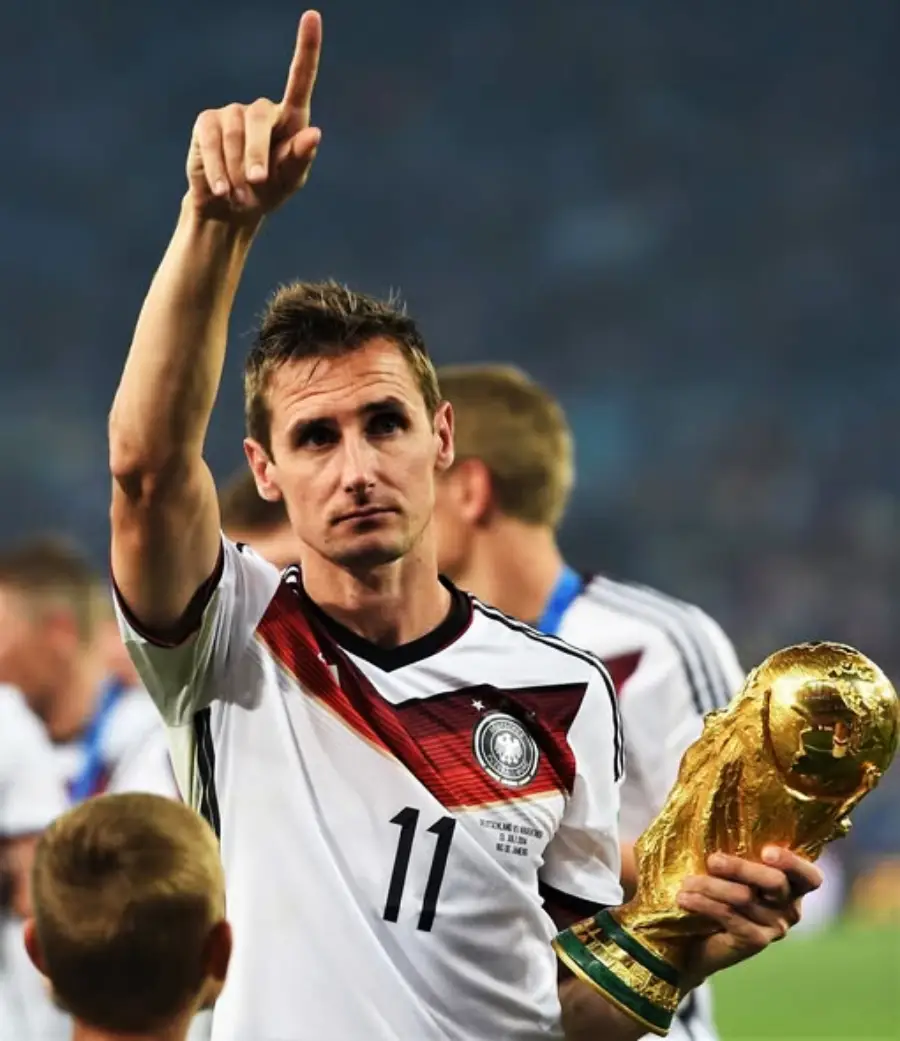 A soccer player in a Germany jersey with number 11 holds the FIFA World Cup trophy in one hand and points upward with the other, celebrating on the field with teammates in the background.