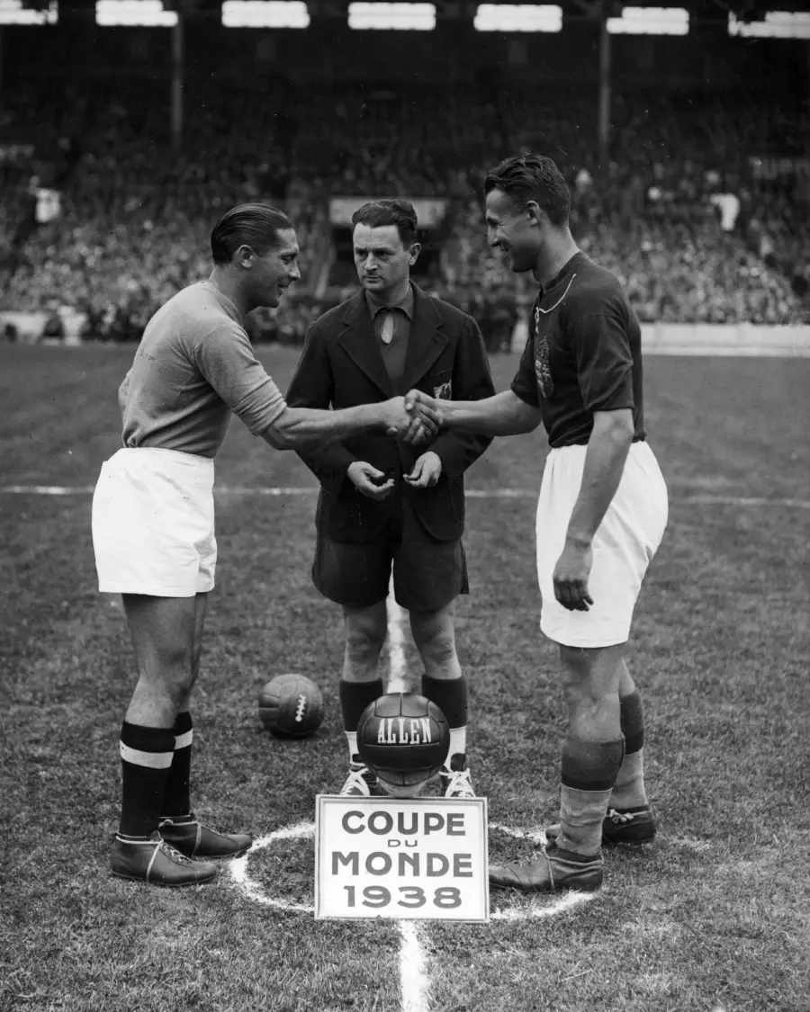 Two football team captains shake hands on the field before a match, with a referee standing between them. A sign reading "Coupe du Monde 1938" and a soccer ball are at their feet, and a stadium crowd fills the background.