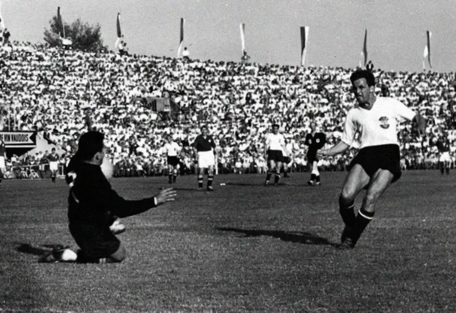 A black-and-white photo of a soccer player in a white jersey taking a shot at goal, while the goalkeeper in dark clothing kneels to block it. A large crowd fills the stadium in the background.