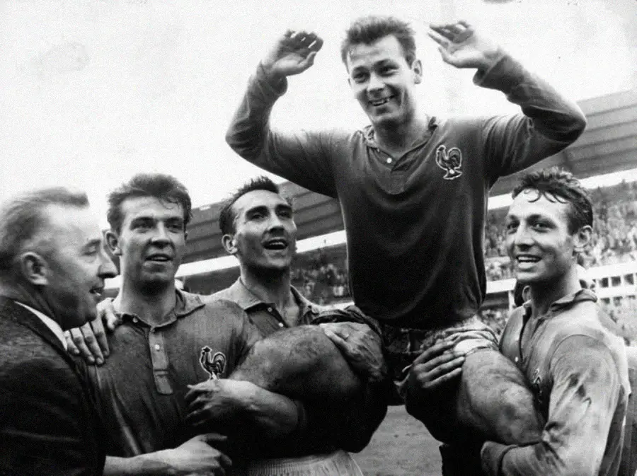 Four soccer players joyfully lift a smiling teammate wearing a French national team jersey, celebrating on the field. A cheering crowd and stadium are visible in the background.