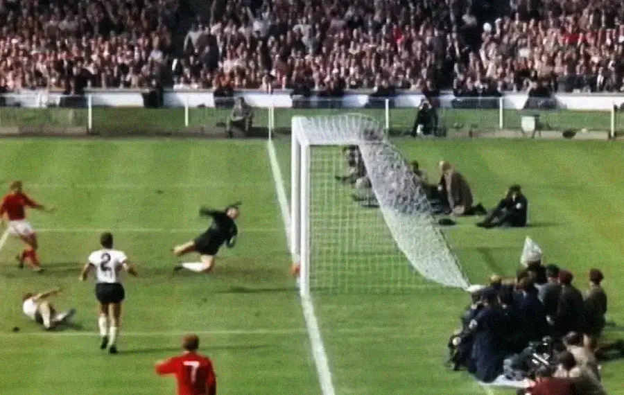 A soccer match scene shows a ball hitting the crossbar and bouncing down near the goal line, with players, a goalkeeper diving, photographers, and a crowd in the stadium.
