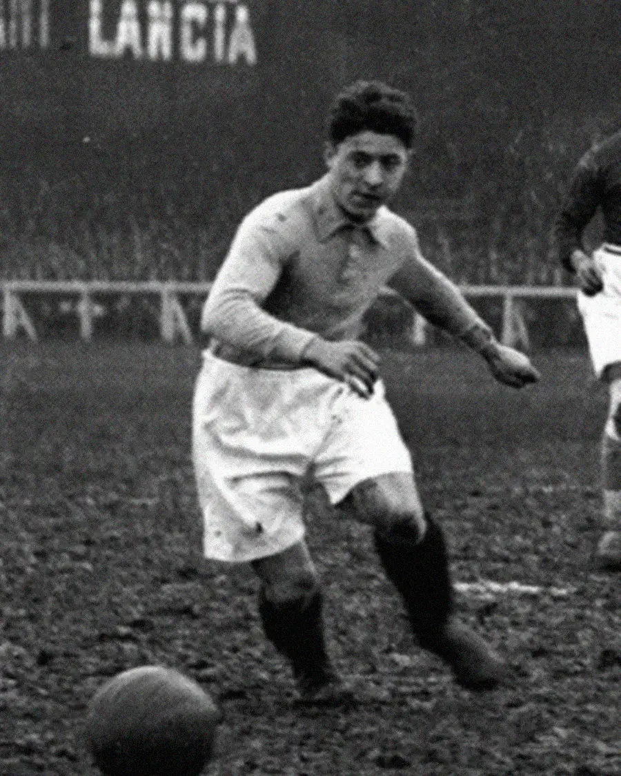 A vintage black-and-white photo of a soccer player in mid-action on a muddy field, wearing a light-colored long-sleeve jersey, white shorts, and dark socks, with an old-fashioned soccer ball in the foreground.