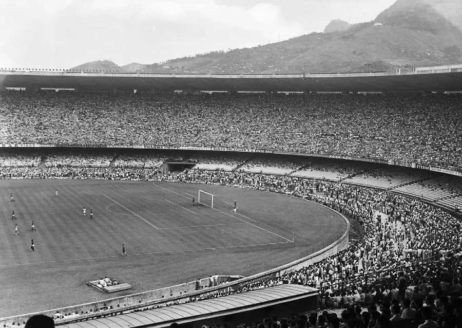 A large, packed soccer stadium with thousands of spectators surrounds a game in progress on the field. Hills and mountains are visible in the background under a cloudy sky.