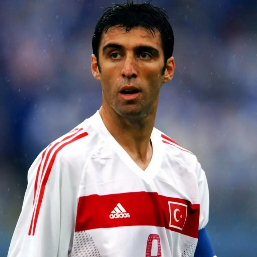 A male soccer player wearing a white and red Turkish national team jersey with the number 9 and a captain's armband stands on the field, looking to the side. The background is blurred.