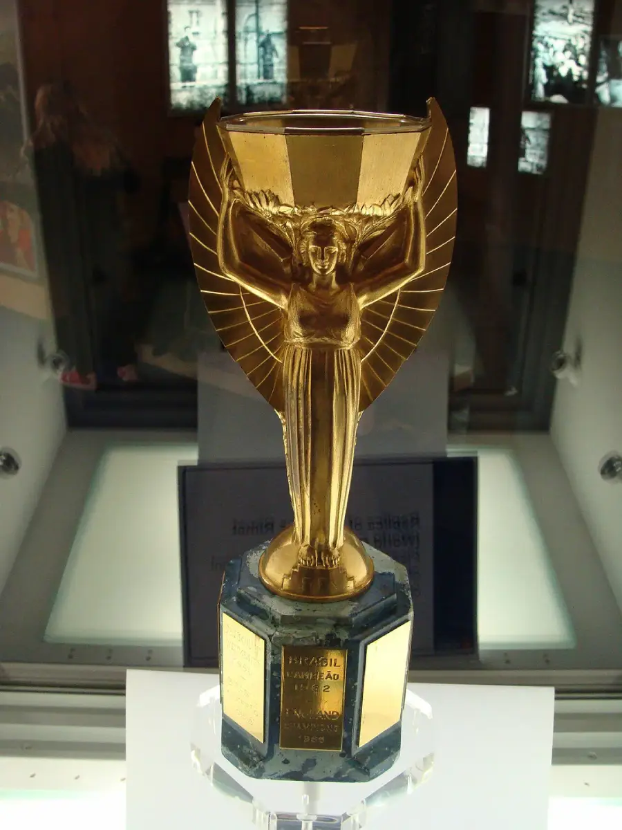 A golden trophy with a winged female figure holding a shallow cup above her head, mounted on a dark, hexagonal base with engraved plaques, displayed in a glass case.