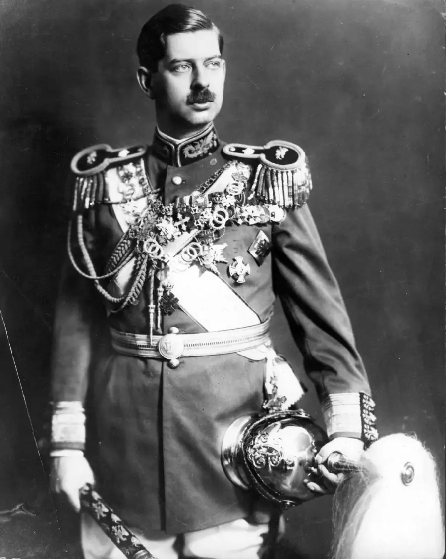 A man in an ornate military uniform adorned with medals and sashes stands holding a helmet. He has a mustache and is looking to the side against a plain dark background.