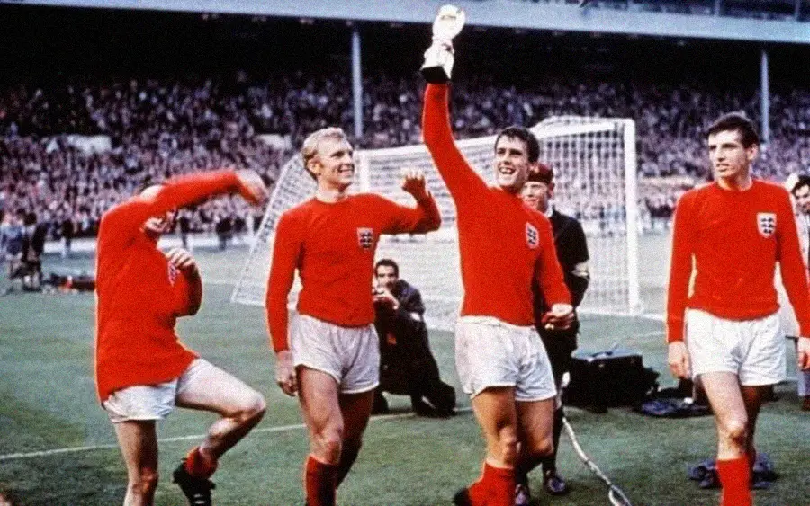 Four soccer players in red England jerseys celebrate on the field, one holding a trophy aloft. A crowd and a goalpost are visible in the background.