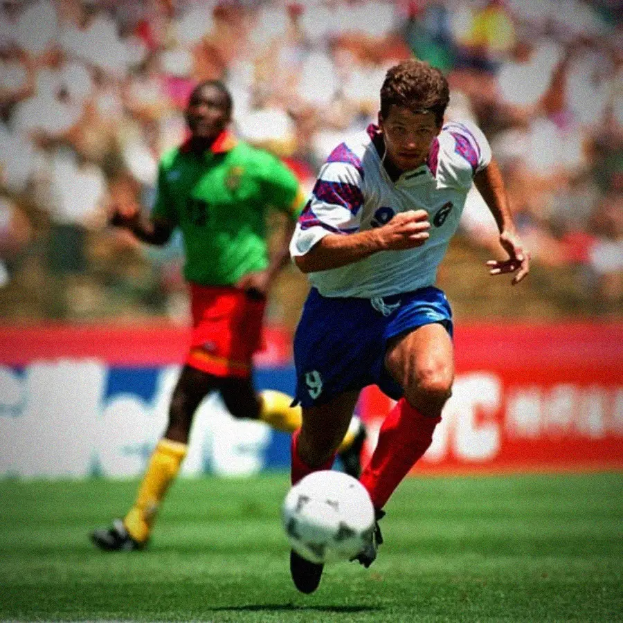 A soccer player in a white and blue uniform runs toward a ball on a field, with another player in a green and red uniform trailing behind during a match. The background shows a blurred crowd in the stadium.