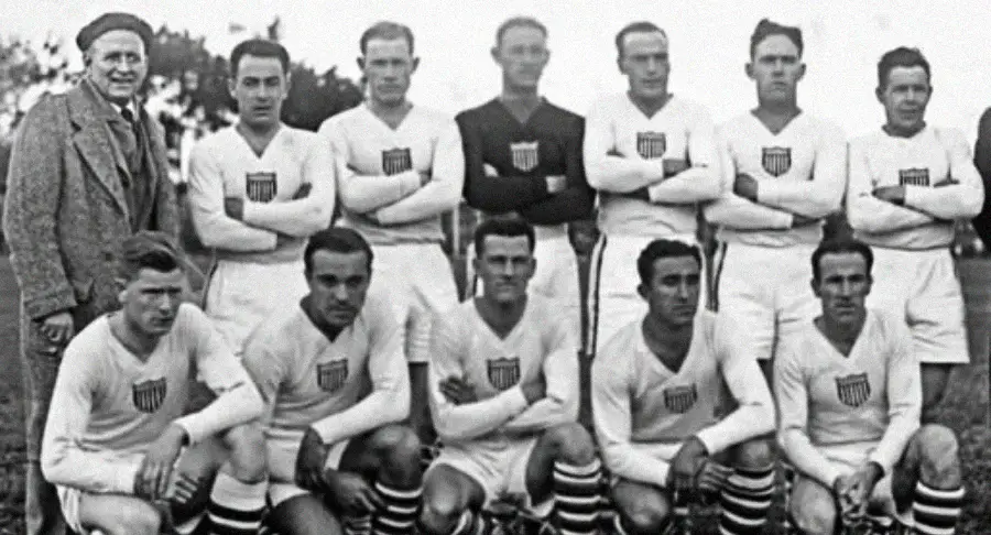 A vintage black-and-white photo of a men's soccer team in white uniforms with USA badges poses for a group portrait; some players are standing with arms crossed, while others are kneeling.